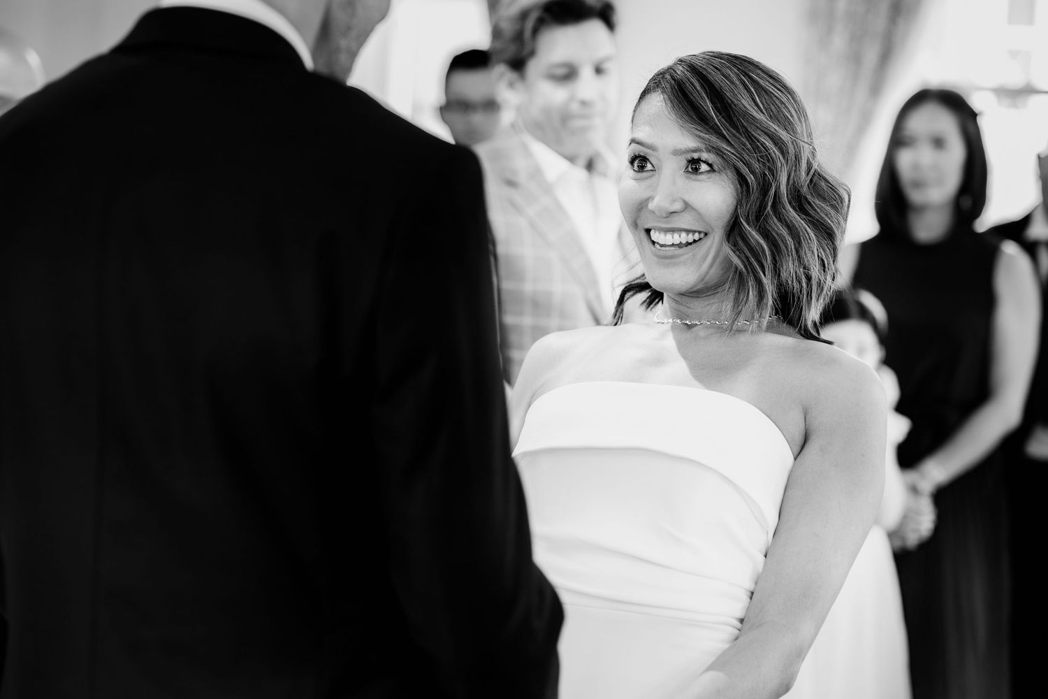 Black and white photo of a bride in a strapless wedding dress smiling during her wedding ceremony.