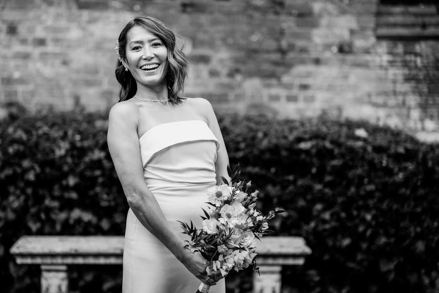A bride in a white wedding dress holds a bouquet while standing against a rustic brick wall background.