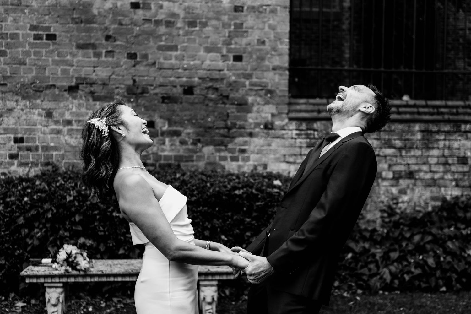 Black and white photo of newlyweds laughing together outdoors against a brick wall with a bouquet nearby.