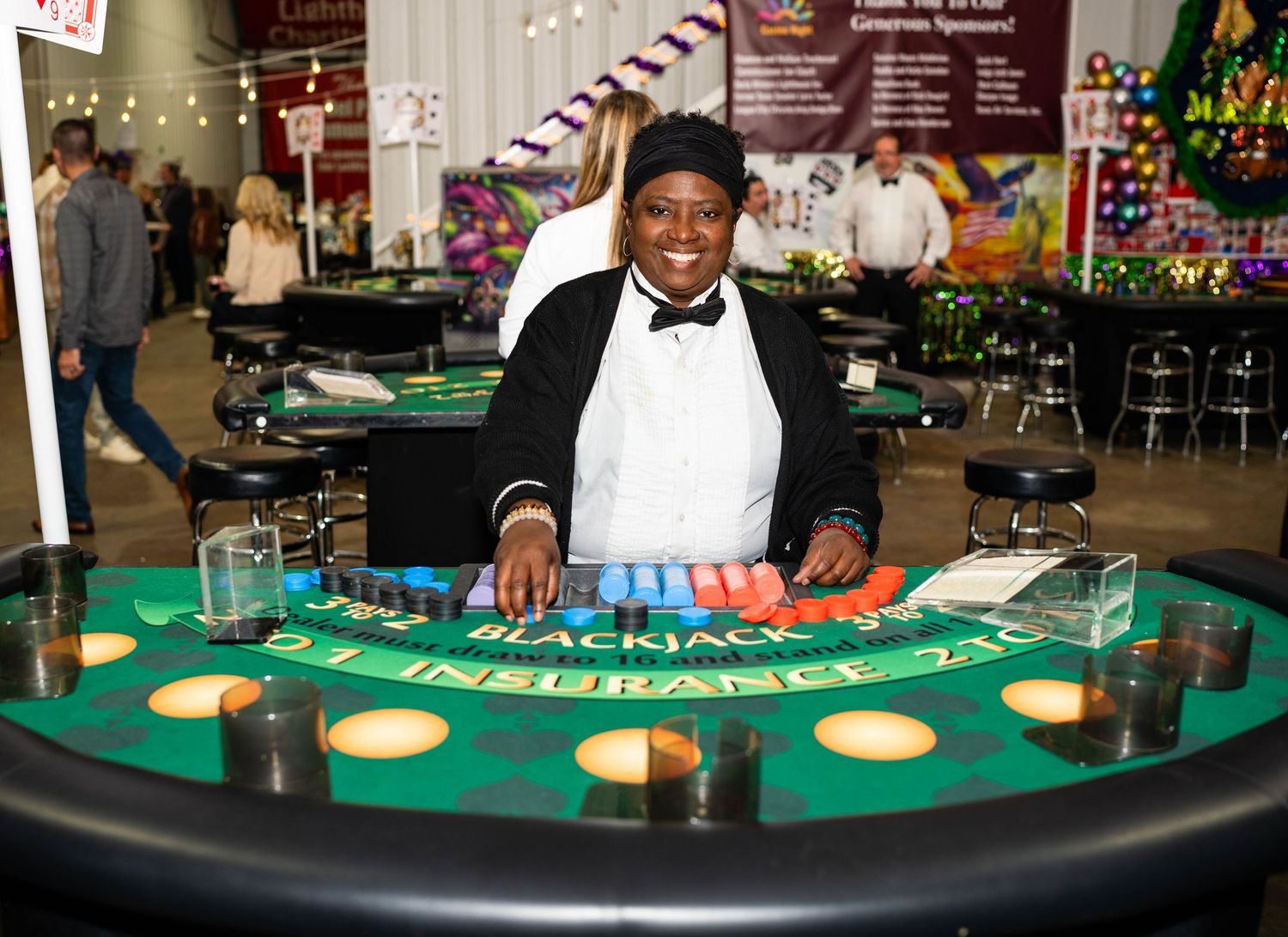 Casino dealers and players interact around a curved blackjack table with green felt surface and betting spots.