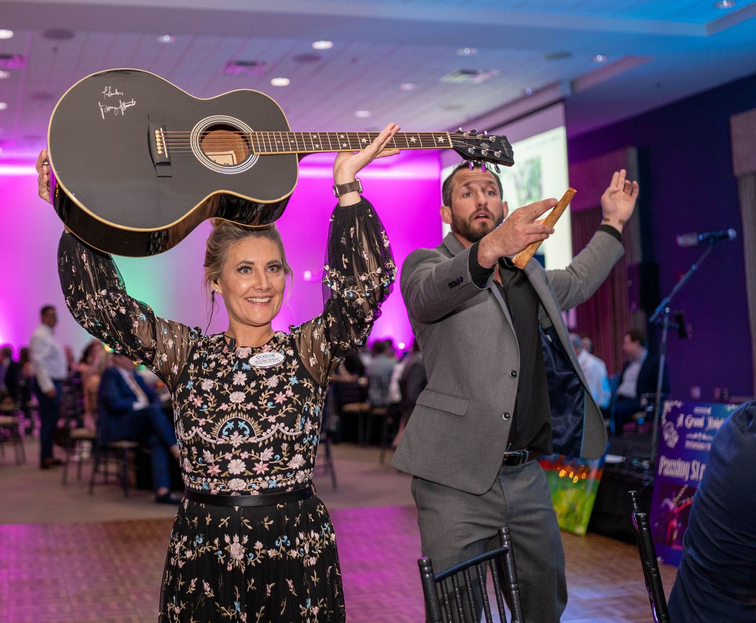 Performers celebrating on stage at an indoor event with purple lighting, one holding an acoustic guitar overhead.