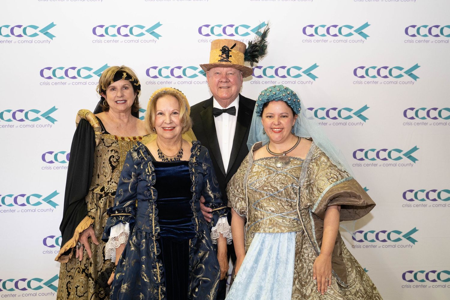 A group poses together at a formal event wearing elegant period costumes and evening wear against a backdrop with logos.