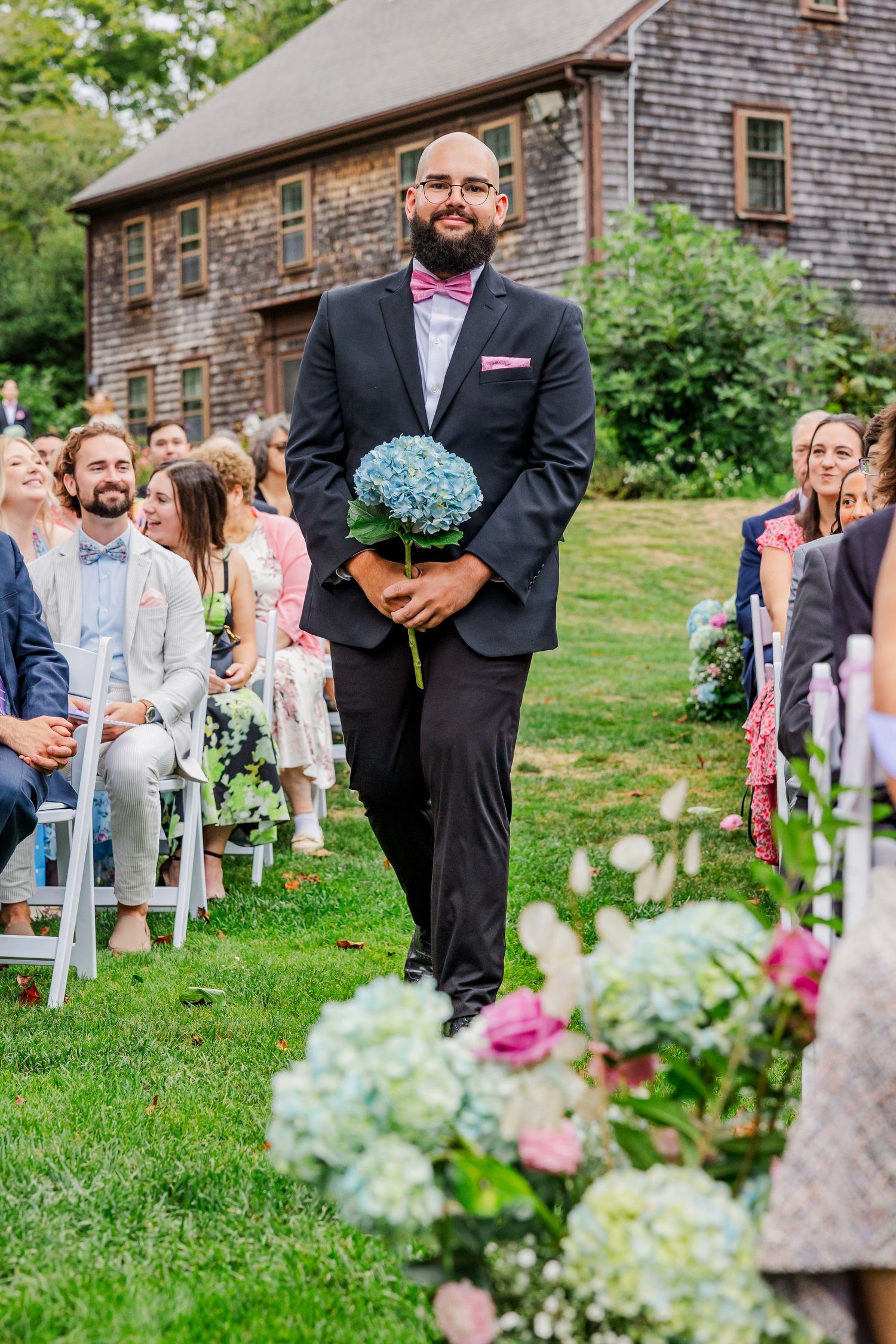 Wedding ceremony guests seated on white chairs on a grassy lawn next to a rustic barn with floral arrangements.