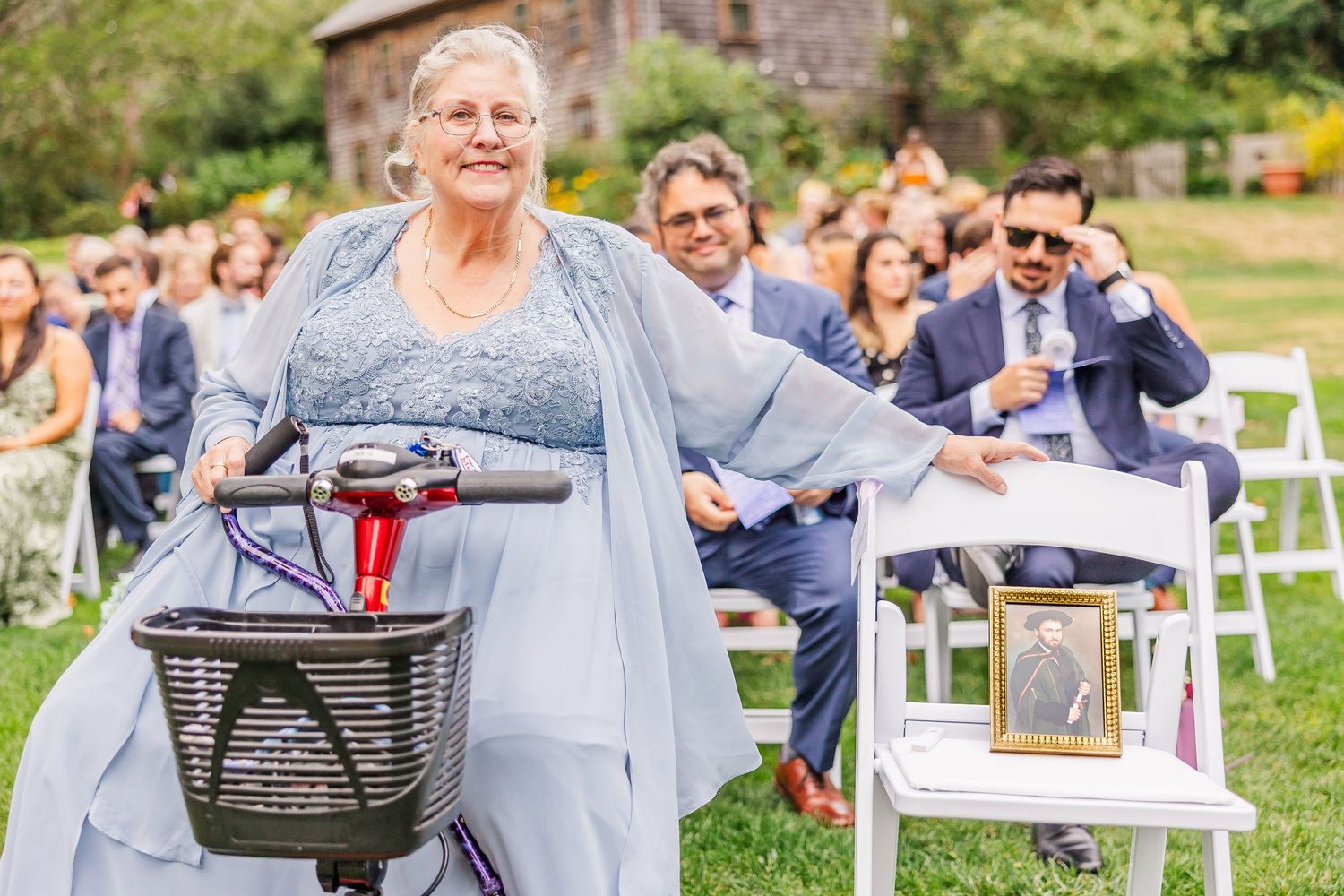 A person in a flowing gray dress rides a mobility scooter at an outdoor wedding ceremony while guests smile in the background.