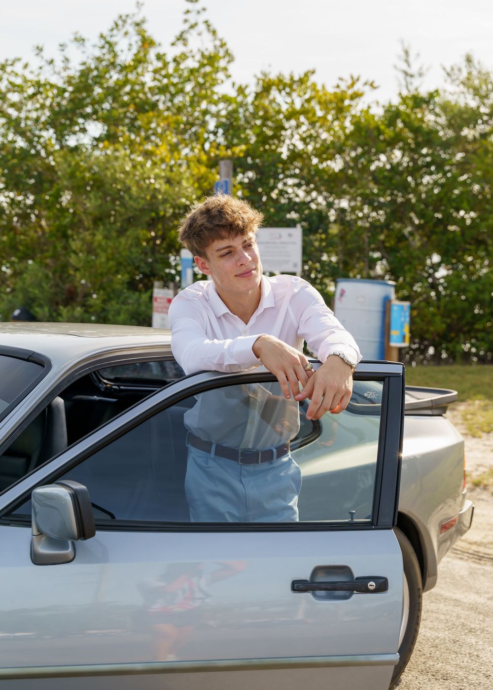 A person in a white shirt leans out of a silver car window in a sunny outdoor parking area.