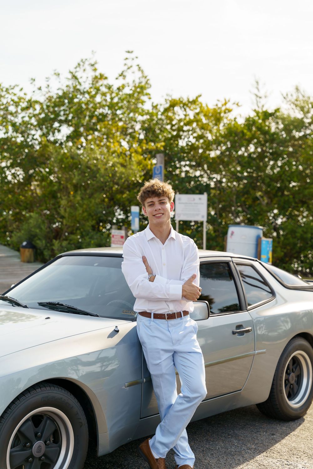 A silver sports car parked outdoors with palm trees and greenery in the background during golden hour.