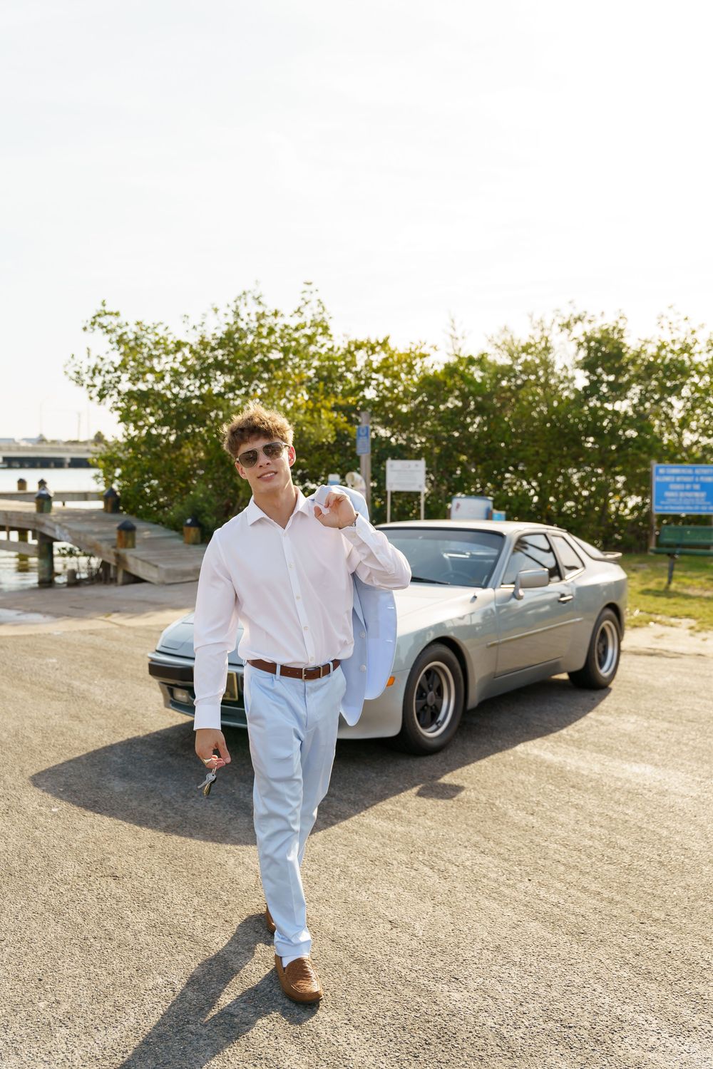 A person in light colored clothing poses next to a classic silver car in a parking lot on a sunny day.