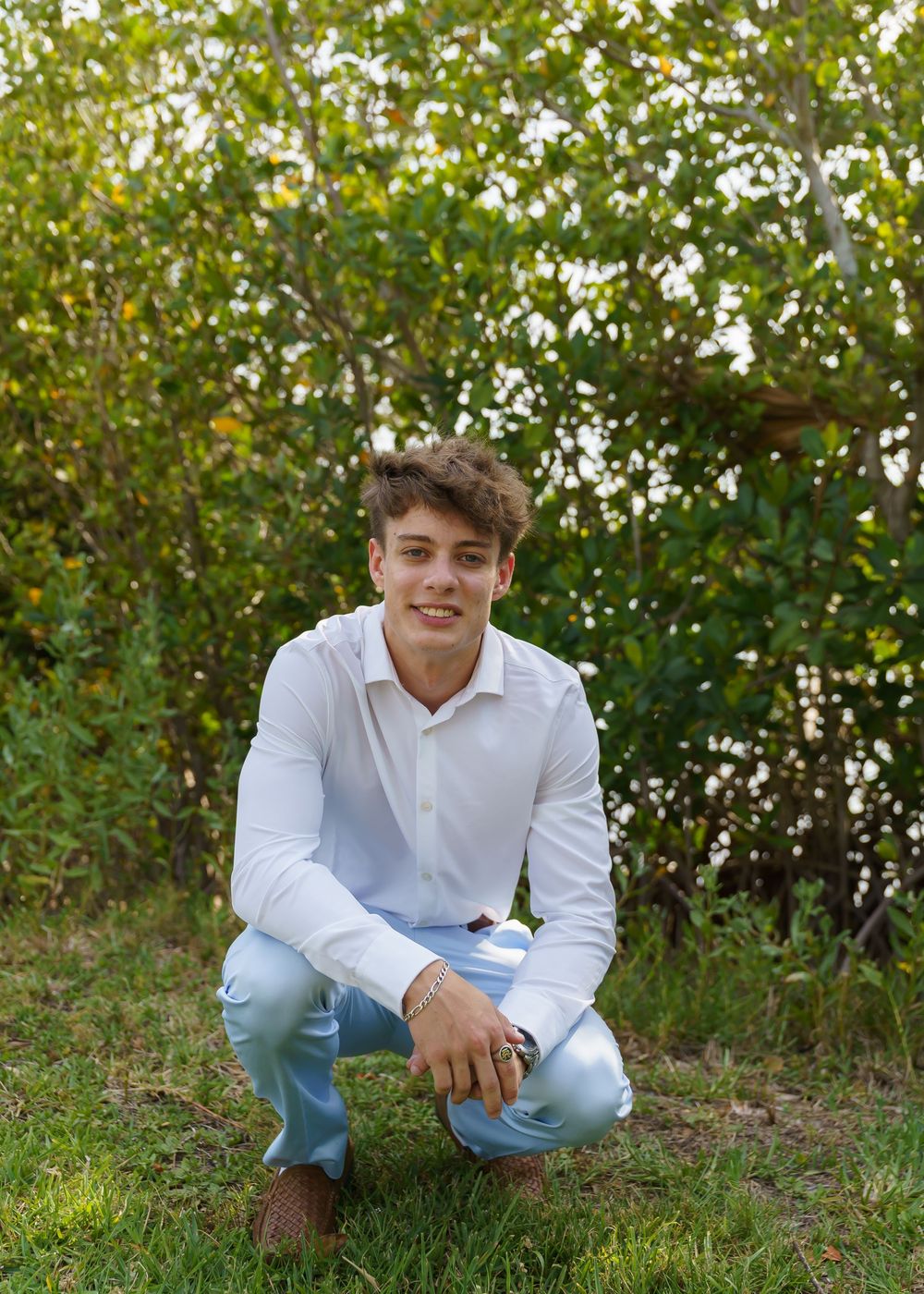 A person in a white shirt and light jeans poses casually in a natural outdoor setting with greenery in the background.