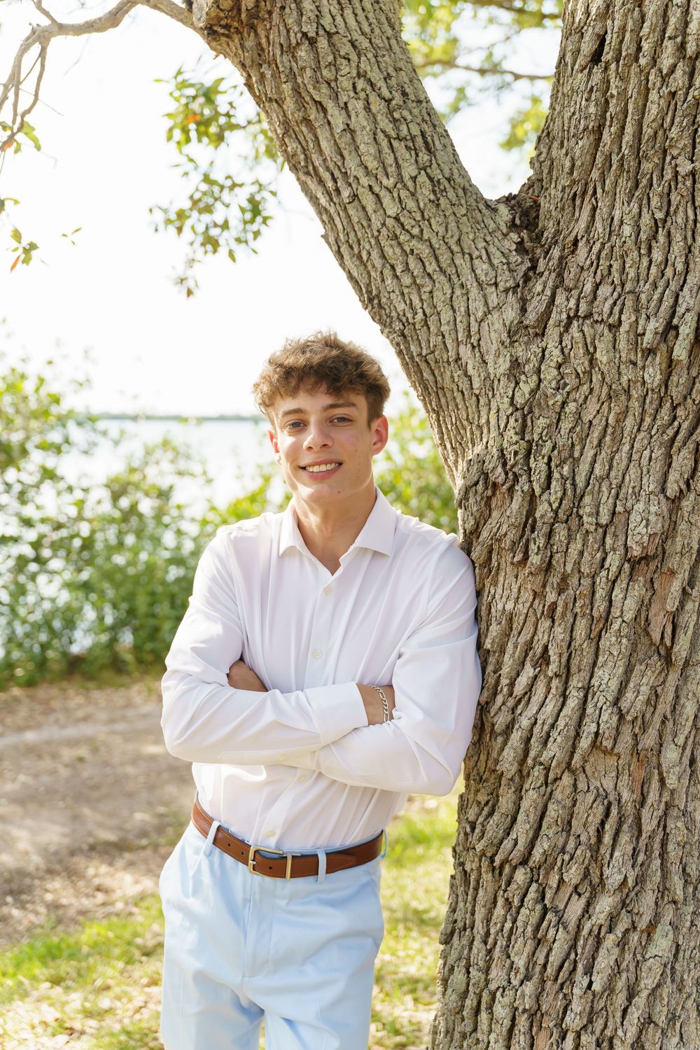 A person in a white button-down shirt and light pants stands with arms crossed by a large tree trunk outdoors.