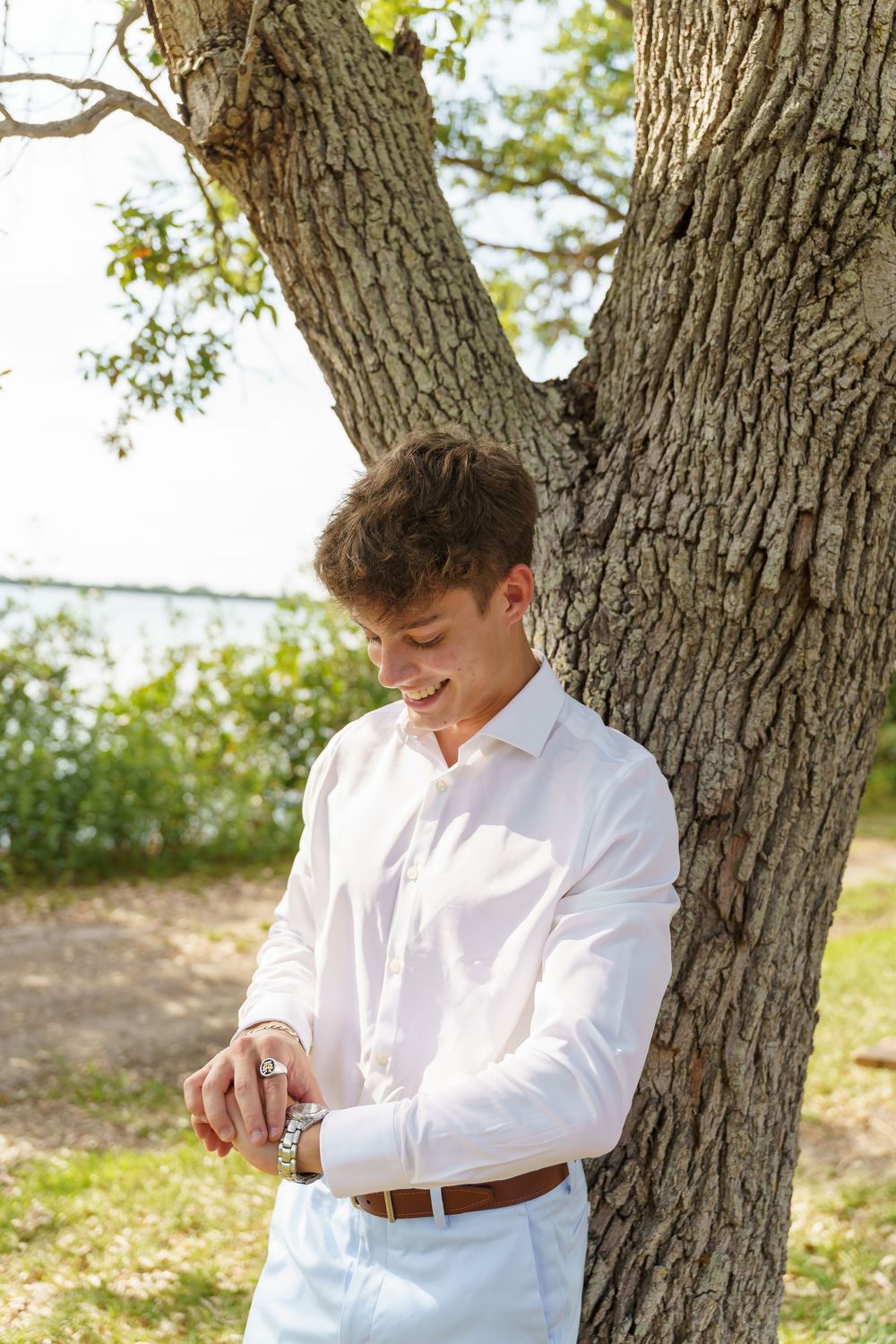 A person in a white button-down shirt and brown belt stands against a tree trunk in natural outdoor lighting.