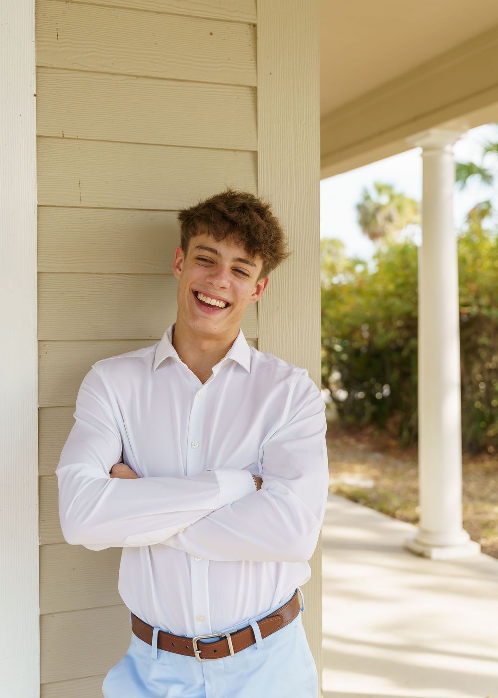 Young person in a white button-down shirt and brown belt poses against white columns in outdoor setting.