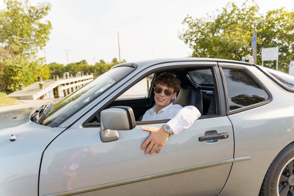 A silver car drives along a road with trees and greenery visible in the background on a sunny day.