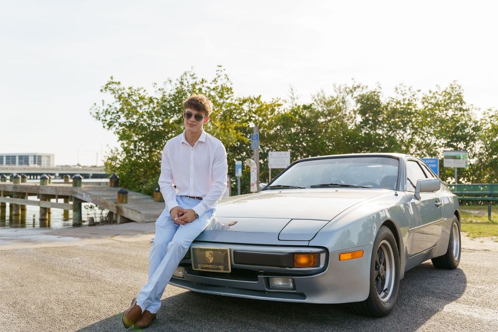 A vintage gray Porsche 944 parked near water with someone casually leaning against it.