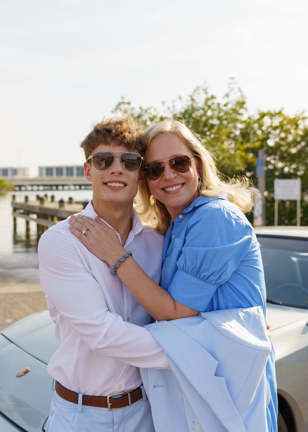 A couple in casual attire embraces and smiles while standing next to a car on a sunny day.