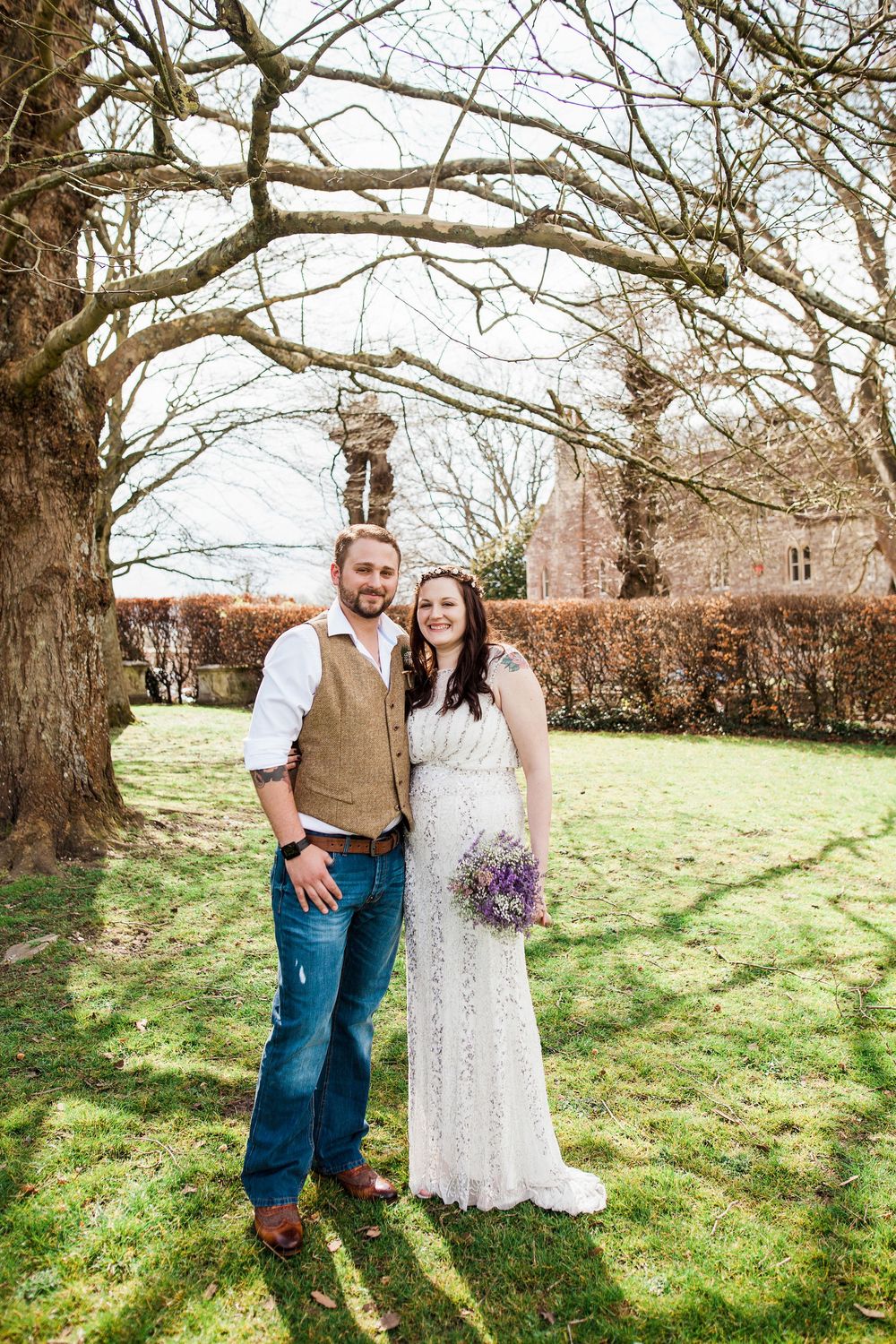 A couple stands together on a grassy hillside under bare winter trees for a rustic outdoor wedding portrait.