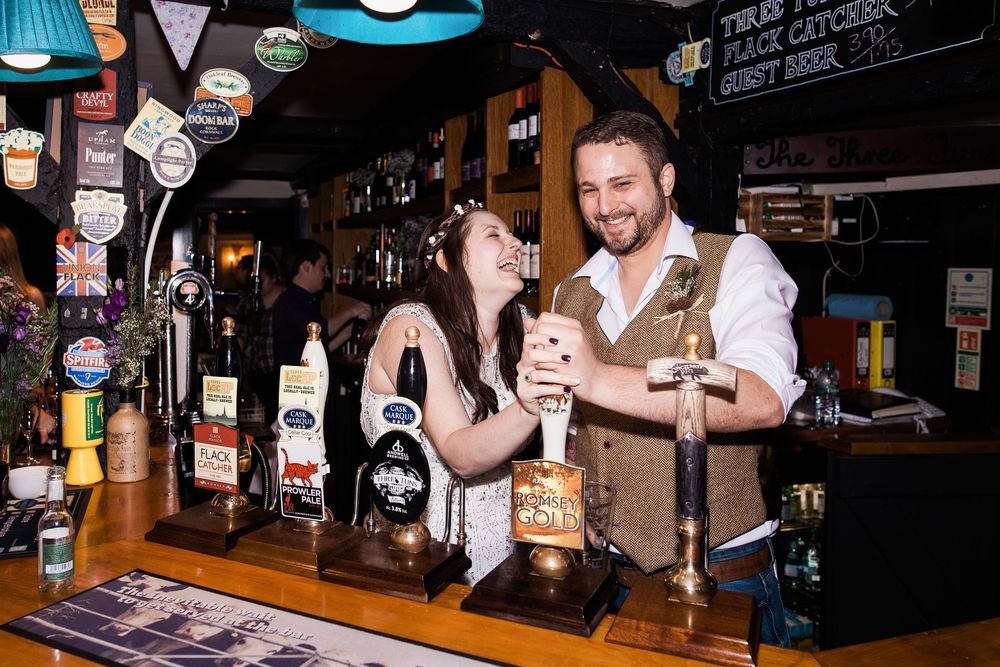 Two bartenders laugh together while working behind a bar counter with various liquor bottles and taps visible.