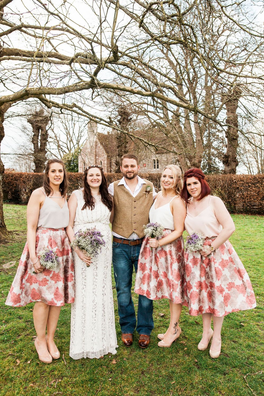Wedding party in floral dresses and rustic attire poses playfully under bare winter trees.