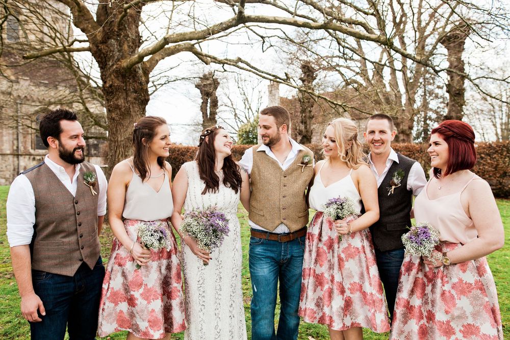 Wedding party poses outdoors in coordinated floral outfits with brown vests and pink dresses against a spring backdrop.