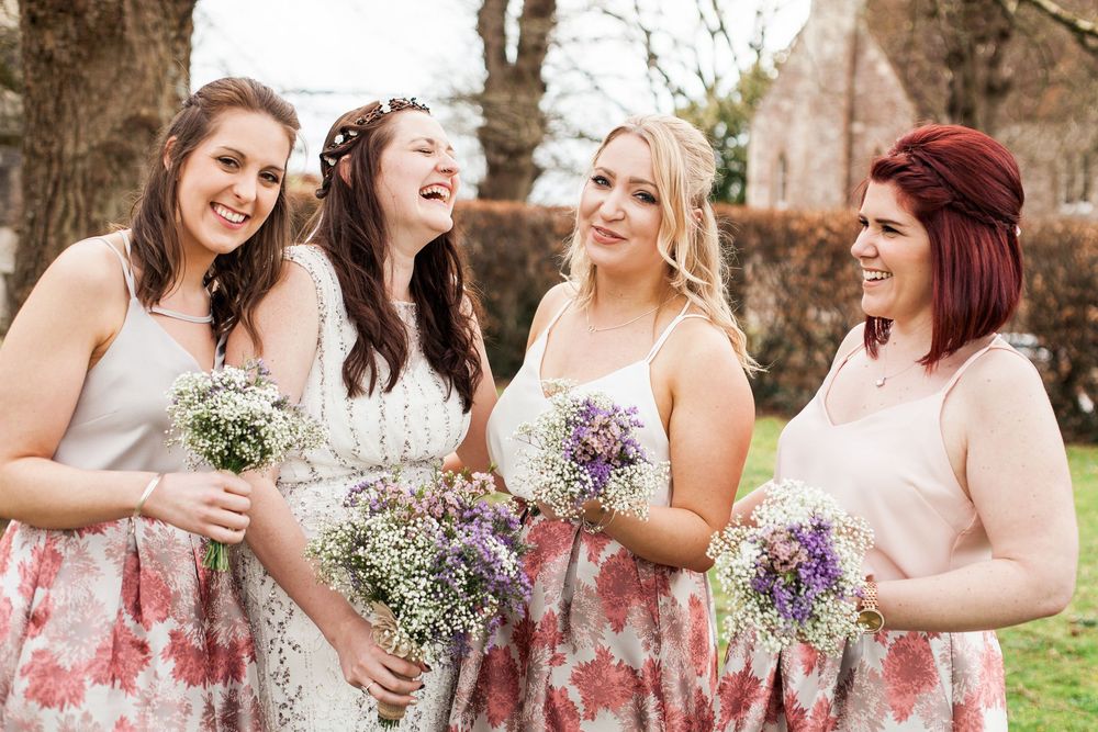 A group of bridesmaids in floral and blush dresses laugh together while holding baby's breath bouquets outdoors.