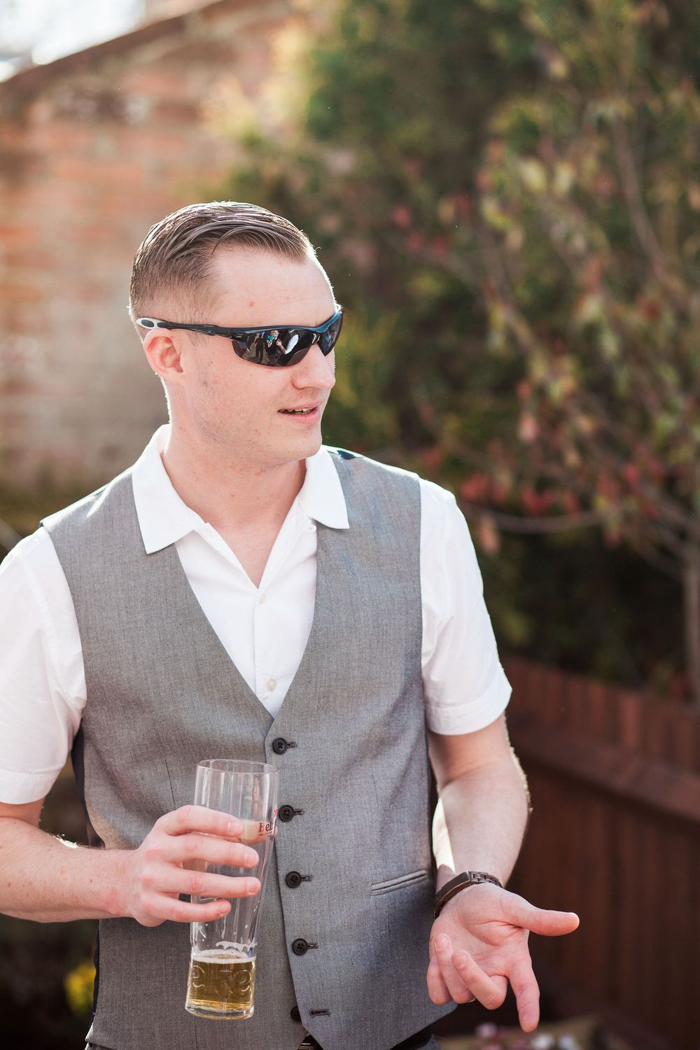 A person in a grey vest and white shirt holds a glass of beer while gesturing outdoors.