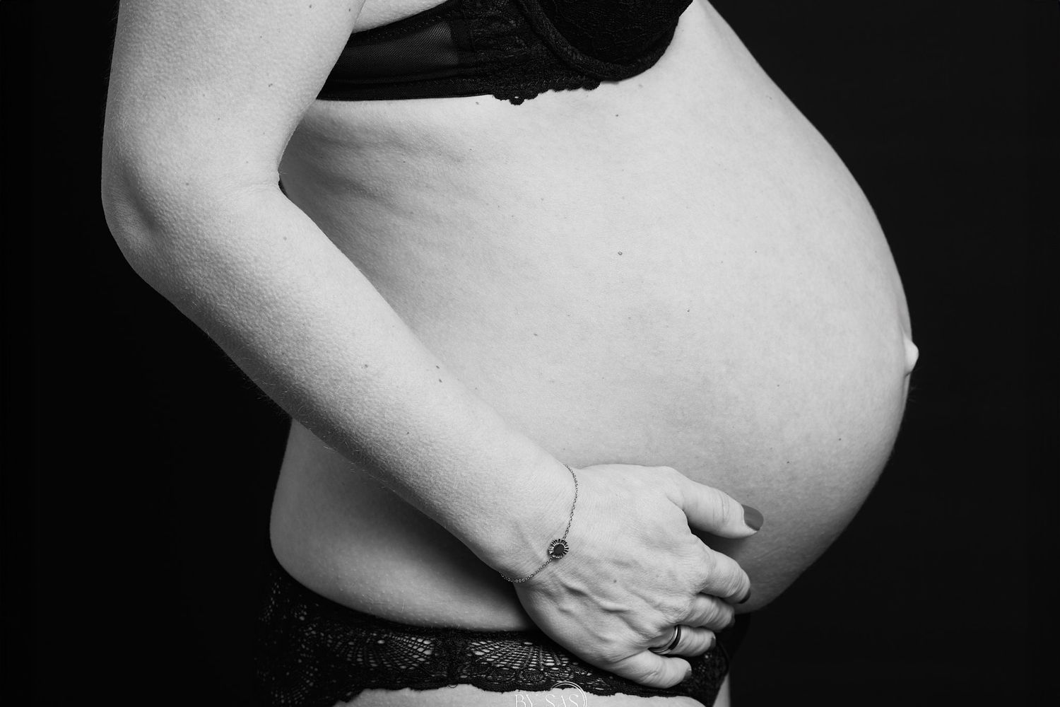 Black and white photo of a pregnant woman cradling her bare baby bump, wearing a dark bra and lace underwear.