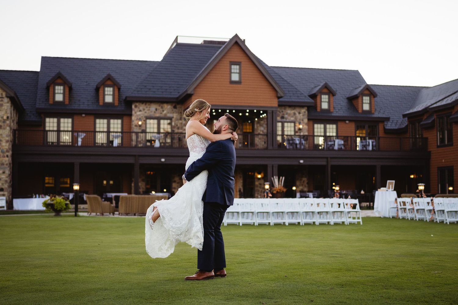 A couple embraces during their wedding celebration on a manicured lawn in front of a grand rustic lodge venue.