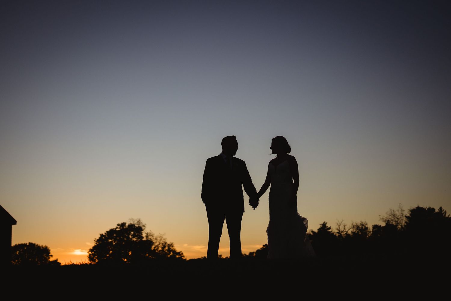 Silhouetted couple holding hands against a vibrant sunset sky with trees and buildings in the background.
