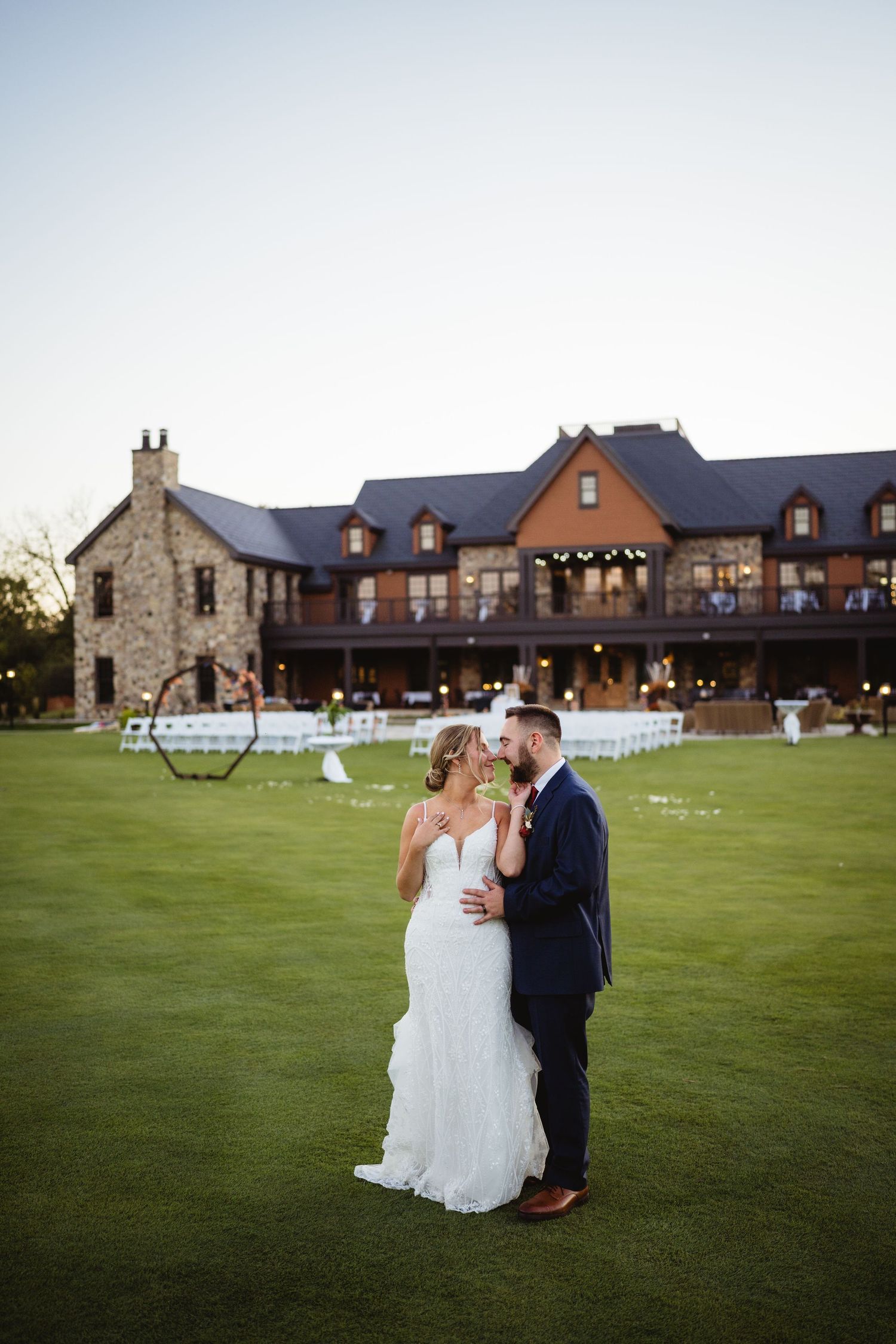 Couple shares romantic moments on a lush green lawn at dusk with rustic stone and brick buildings in the background.