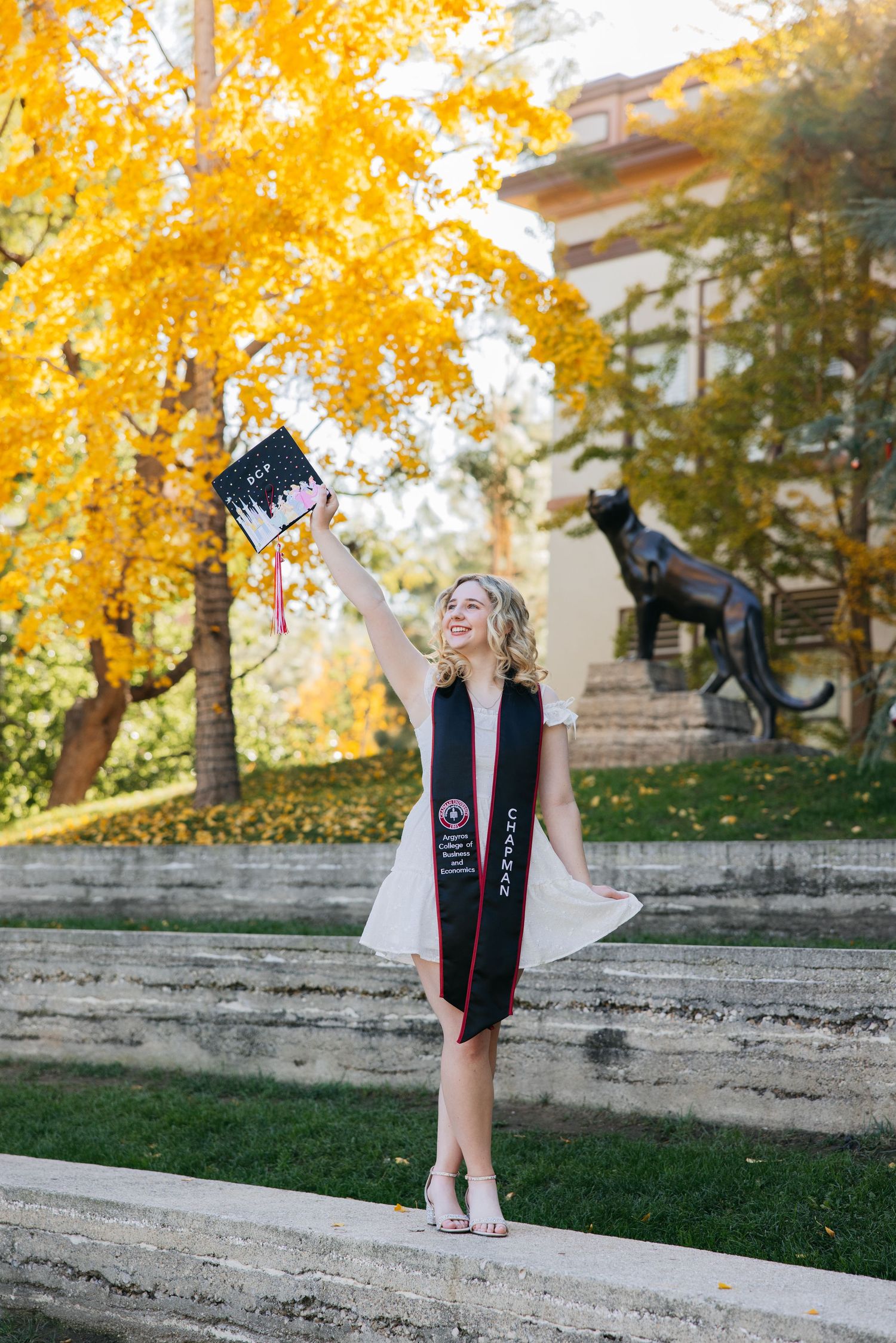 Graduate in cap and gown poses under vibrant yellow fall foliage with bronze statue in background.