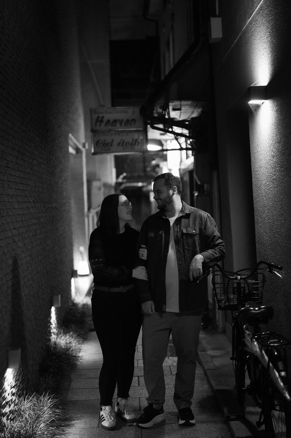 Black and white photo of a couple walking together through a narrow illuminated Japanese alley.