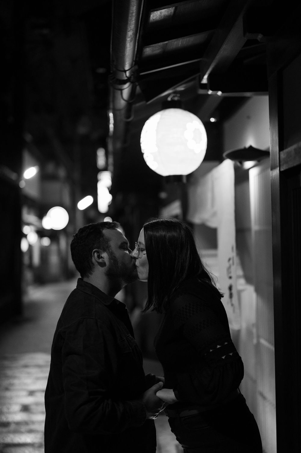 Black and white photo of a couple sharing a kiss beneath a glowing lantern on a night street.