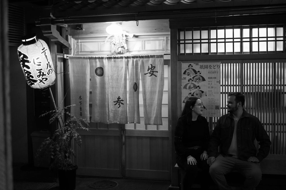 Black and white photo of a couple sitting outside a traditional Japanese restaurant at night.