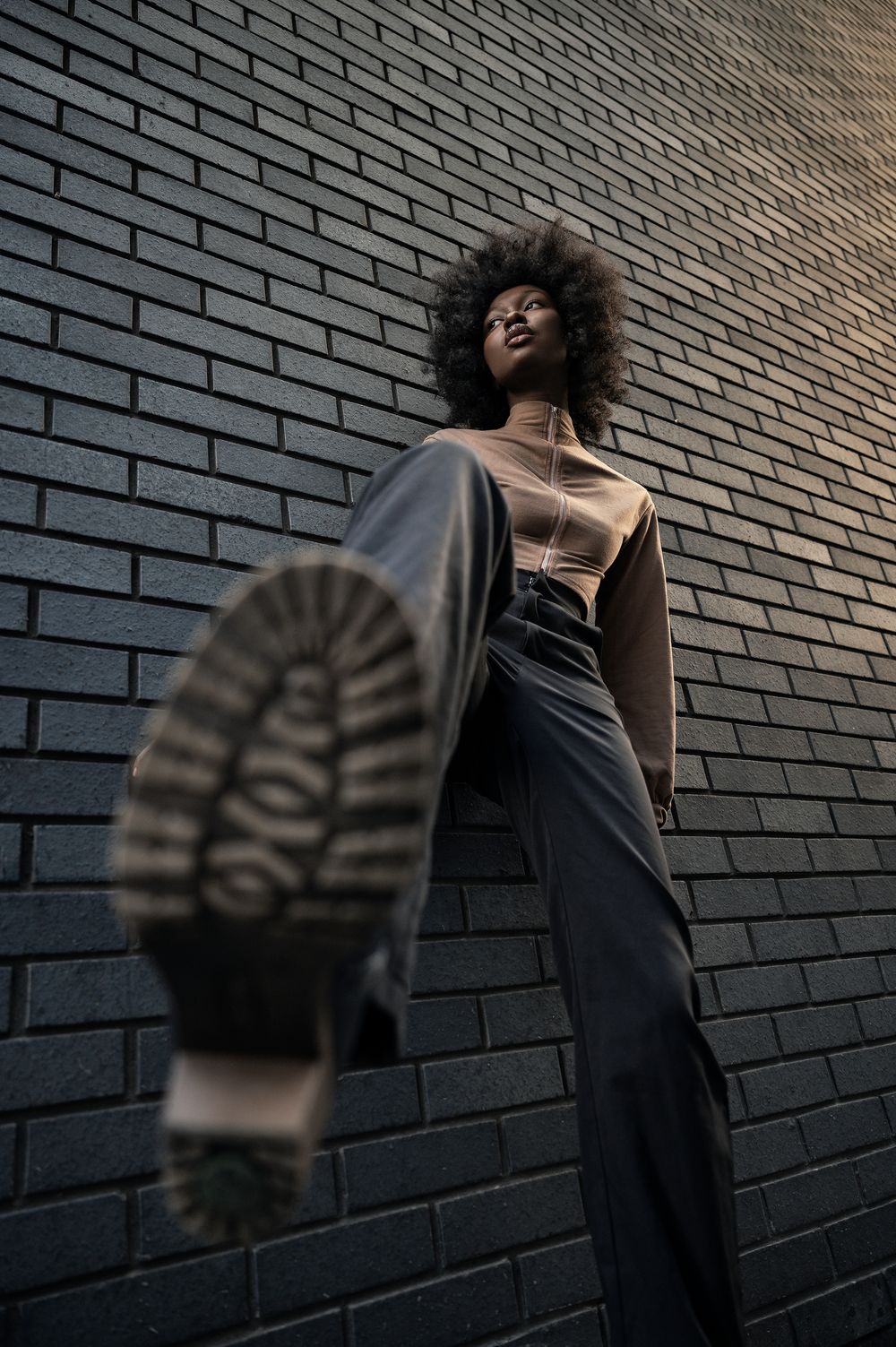 A figure in black clothing poses dramatically against a dark brick wall with artistic shadows and lighting effects.