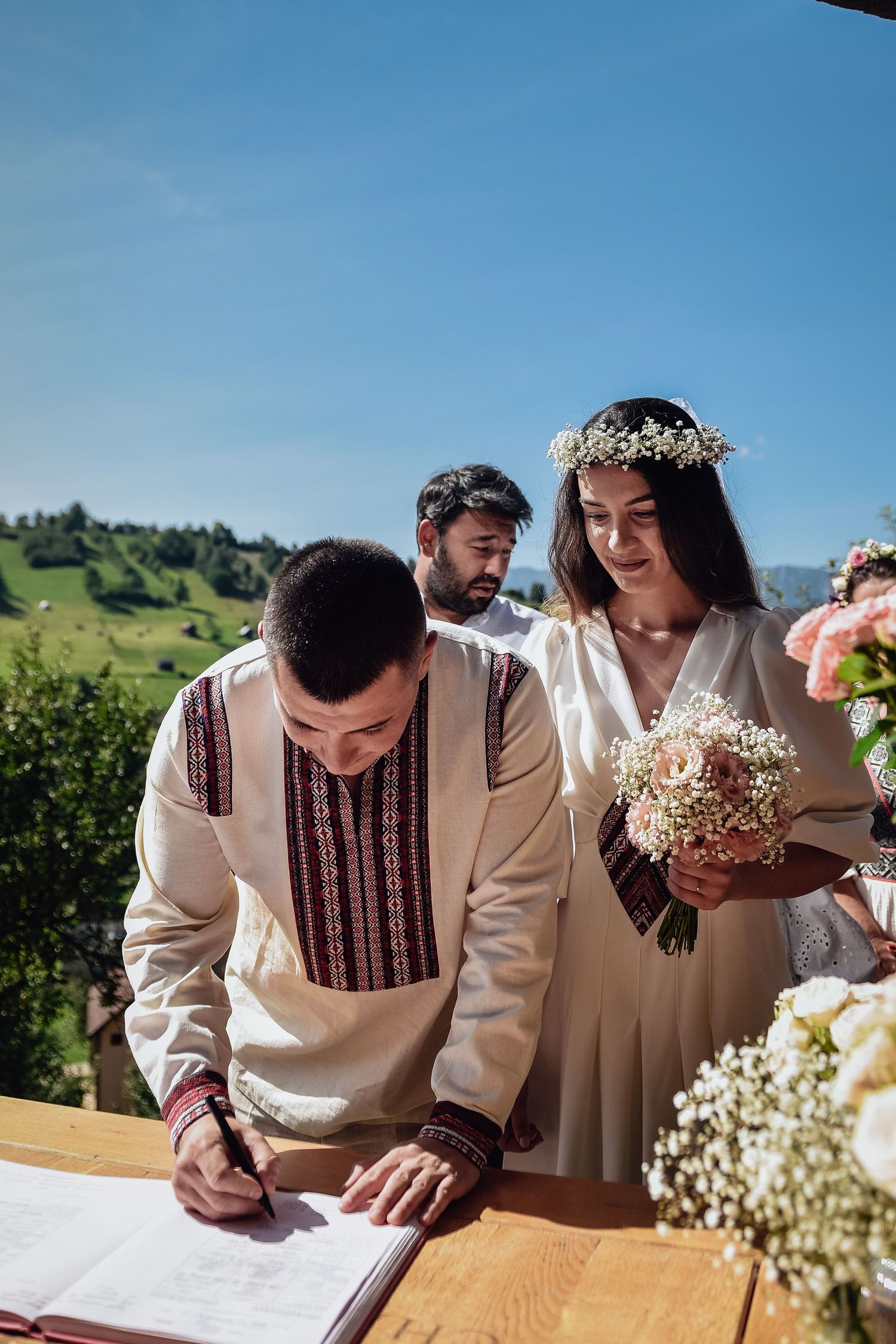 A couple in traditional embroidered wedding attire signs documents at an outdoor ceremony with mountains in the background.