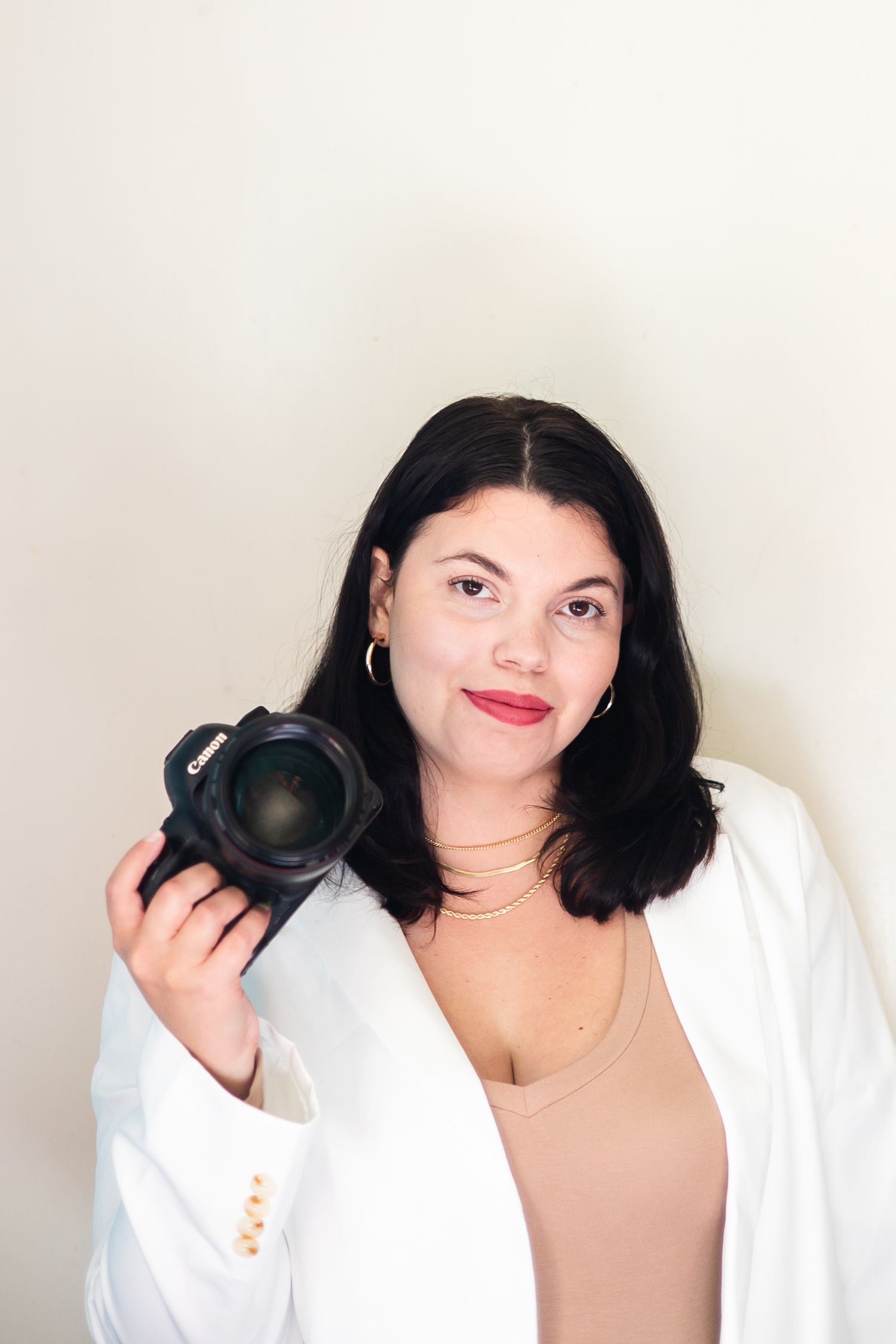 Professional portrait of a photographer in a white  shirt tan belt and beige pants holding a MacBook laptop against a light background.