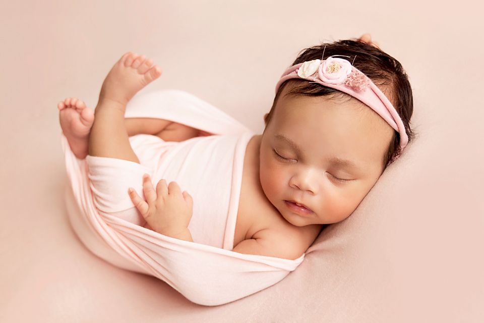 Sleeping newborn wrapped in soft pink fabric with matching headband rests peacefully on pink background.