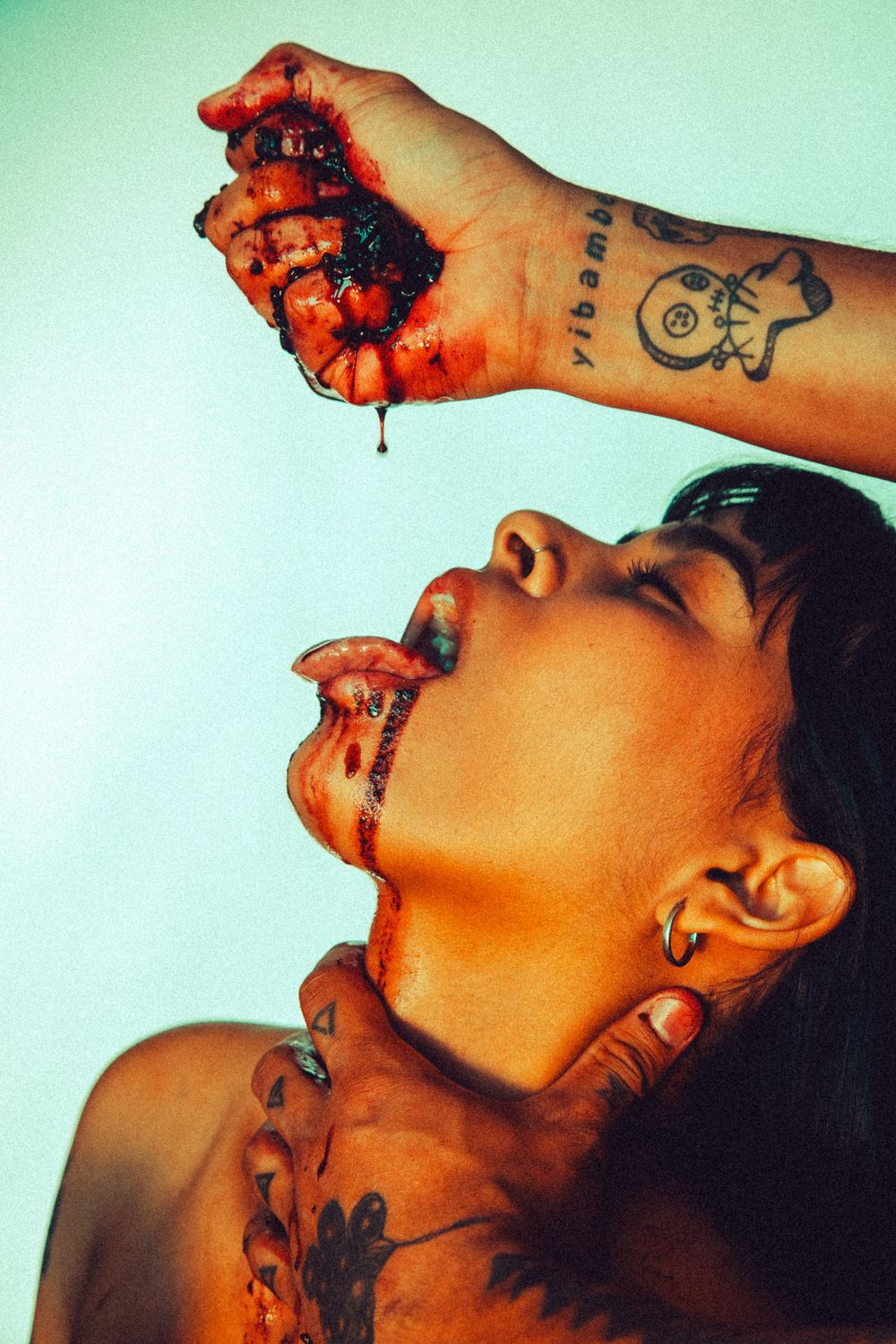 Tattooed woman with mouth open catching dripping red juice squeezed from fruit held above her head.
