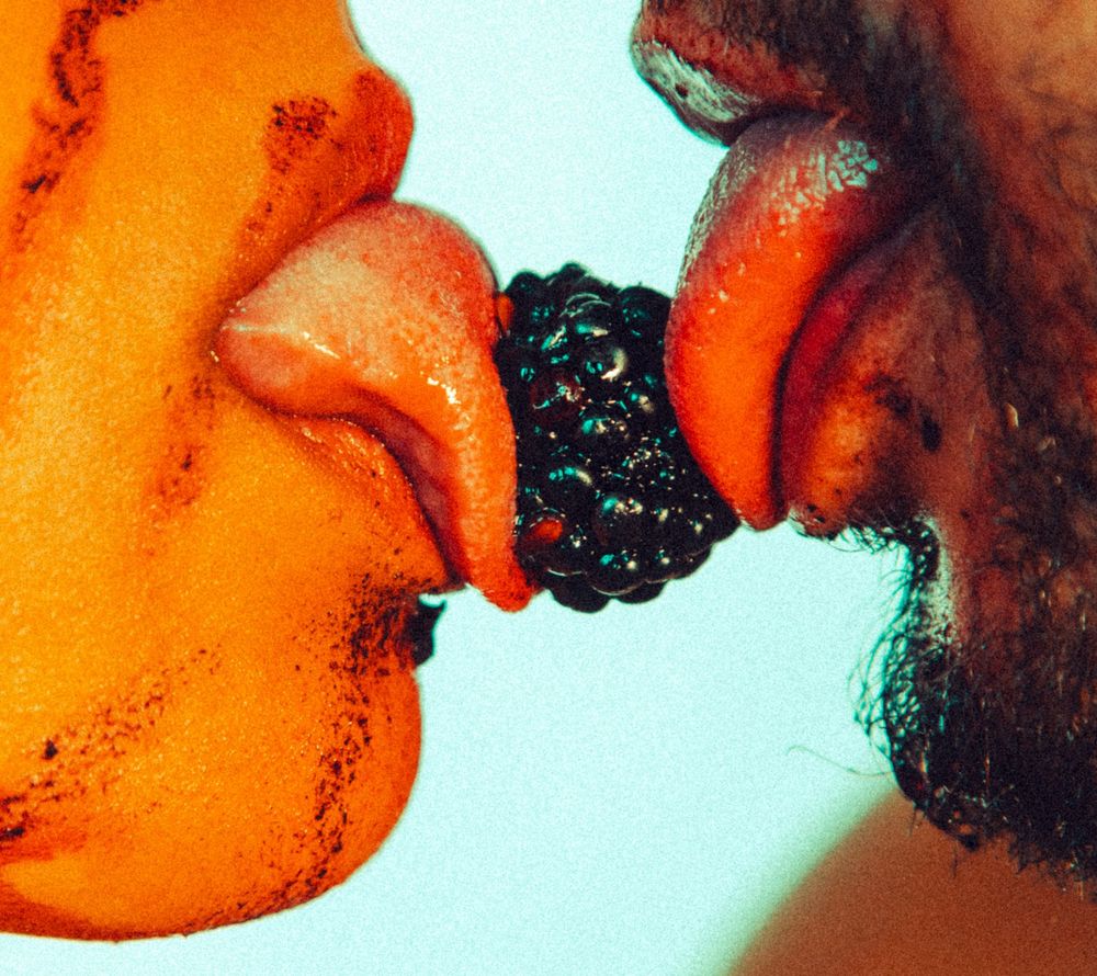 Two people sharing a blackberry with their tongues touching the fruit from opposite sides.