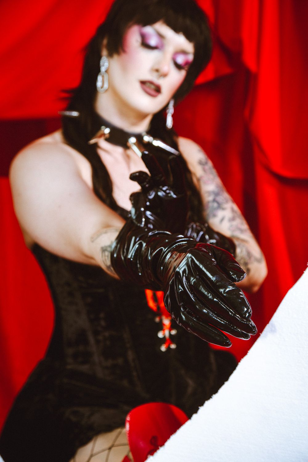 Woman in black corset and shiny latex gloves poses dramatically against red curtain backdrop.