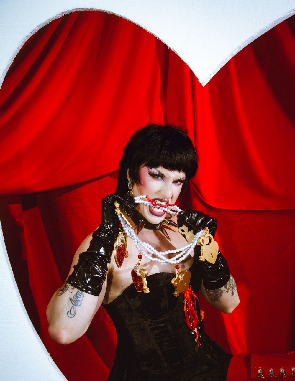 Tattooed woman in black corset and vinyl gloves poses with pearl necklace before a red heart backdrop.
