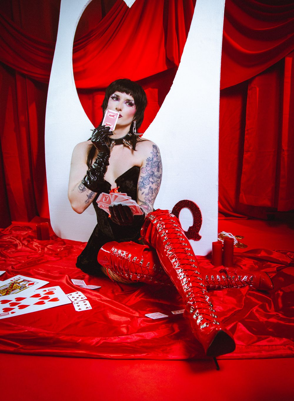 Tattooed woman in black corset and red thigh-high boots sits among playing cards before red curtains.