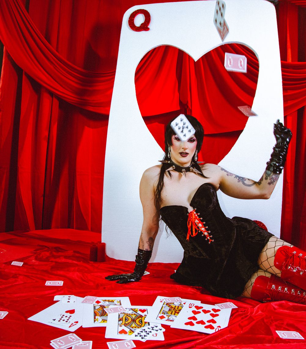 Queen of Hearts performer poses with playing cards in red velvet burlesque setting.