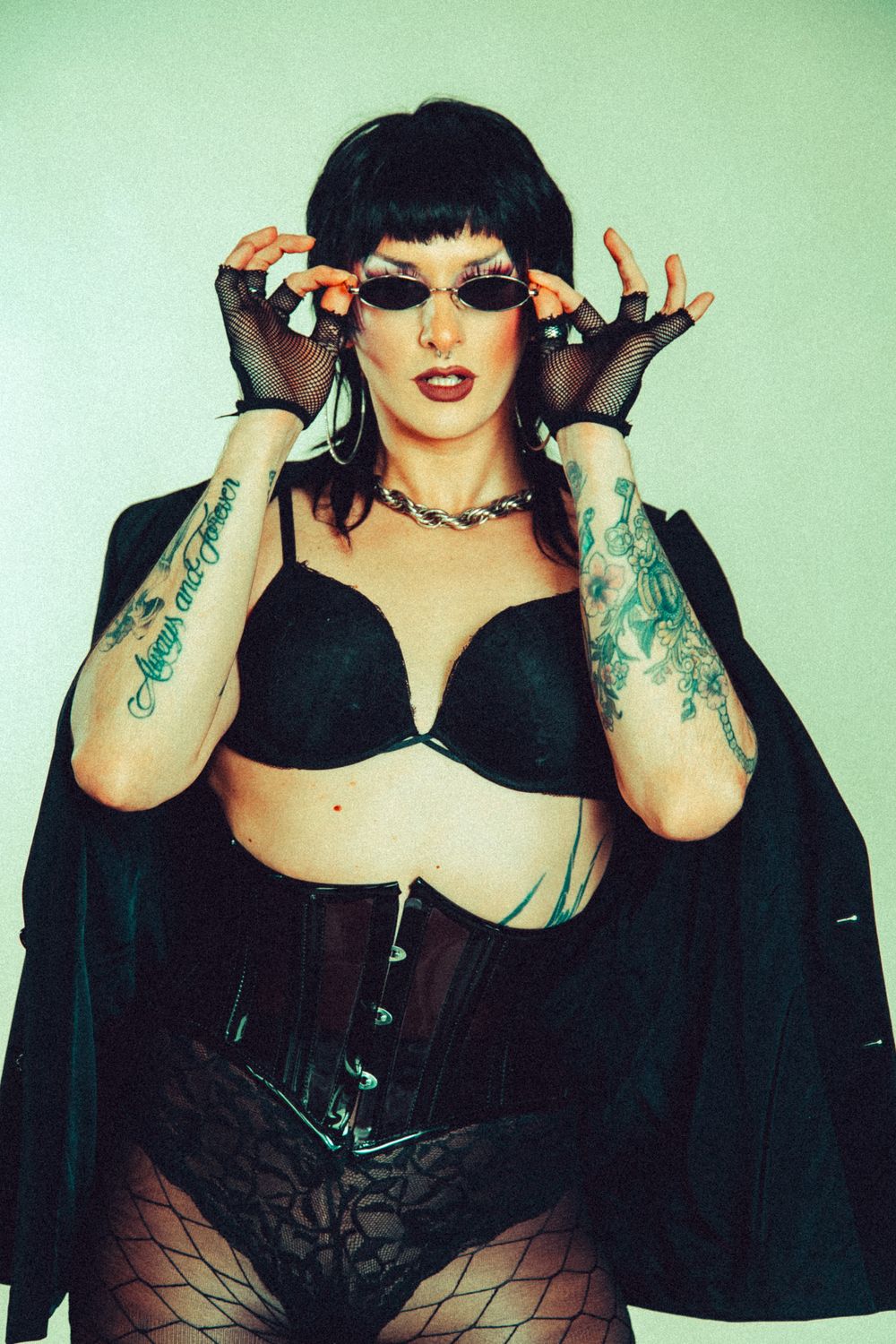 Tattooed woman in black lingerie, corset, and fishnet gloves adjusting sunglasses, wearing a chain necklace.