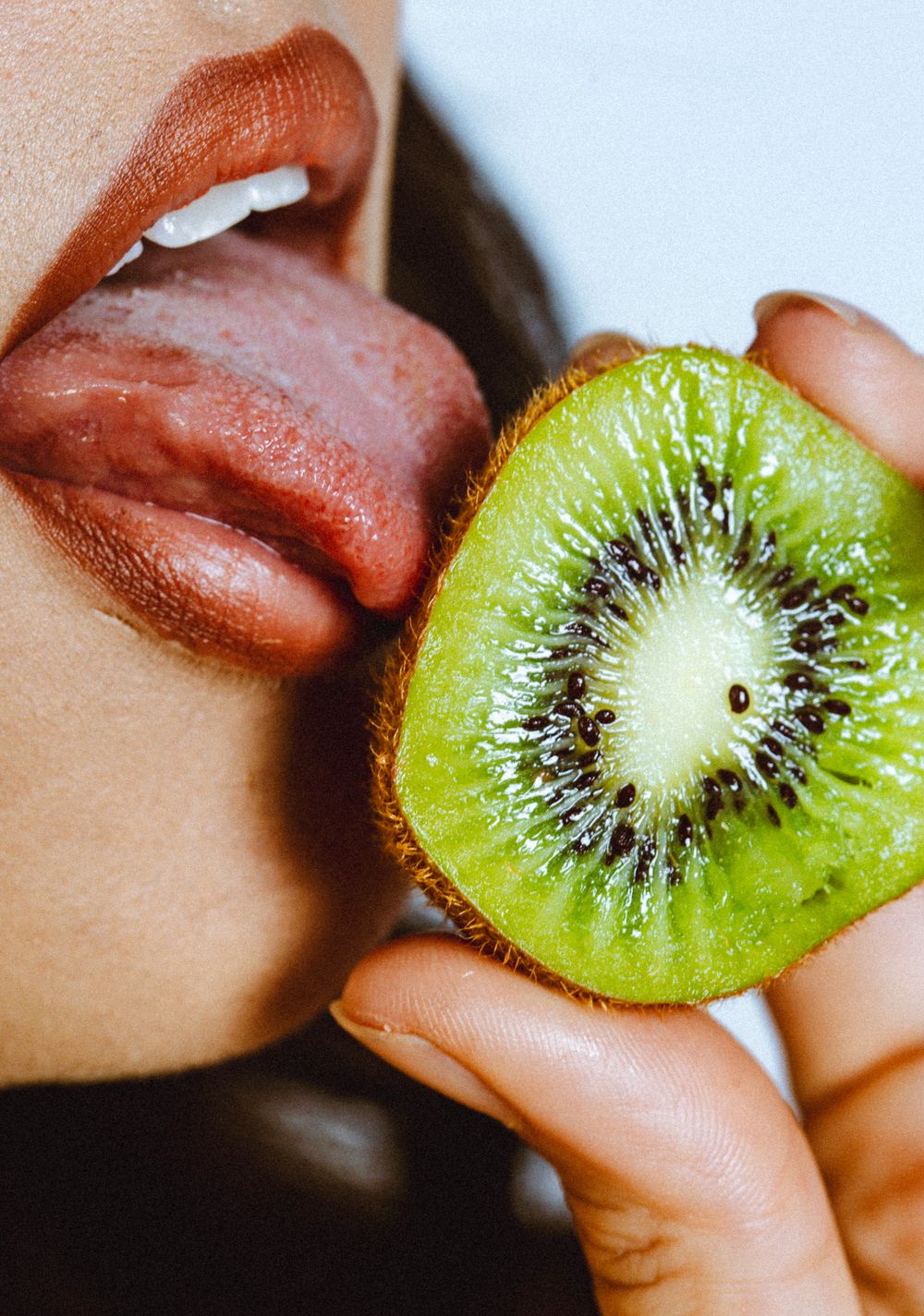 Close-up of a person licking a fresh sliced kiwi fruit, showcasing its vibrant green flesh and black seeds.
