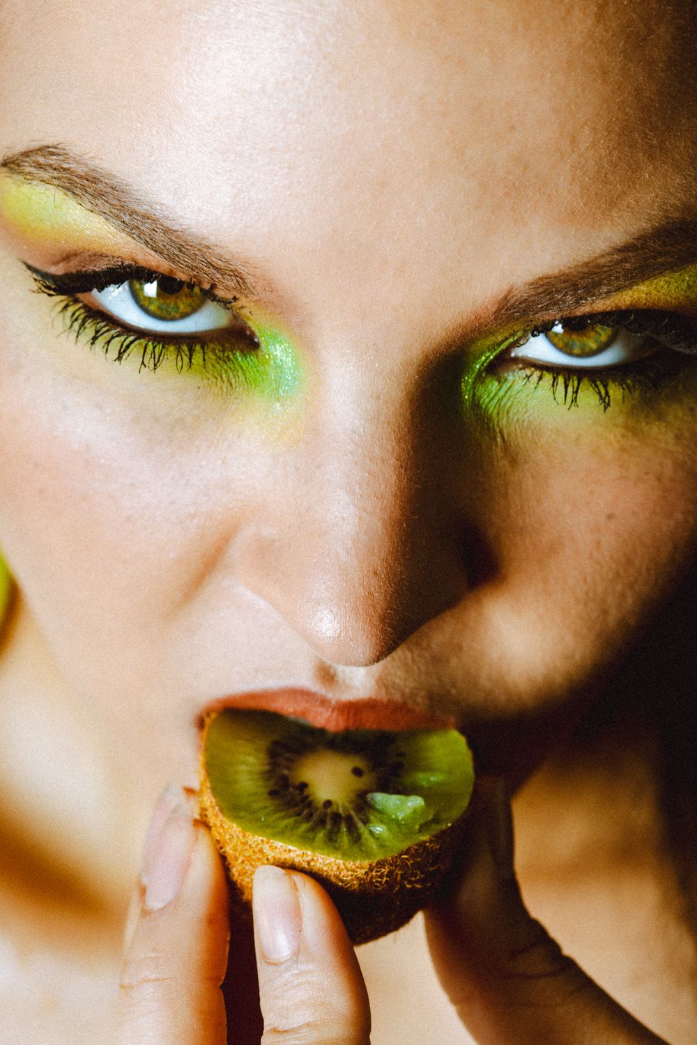 Woman with green eye makeup holding a sliced kiwi fruit to her lips in a close-up beauty shot.