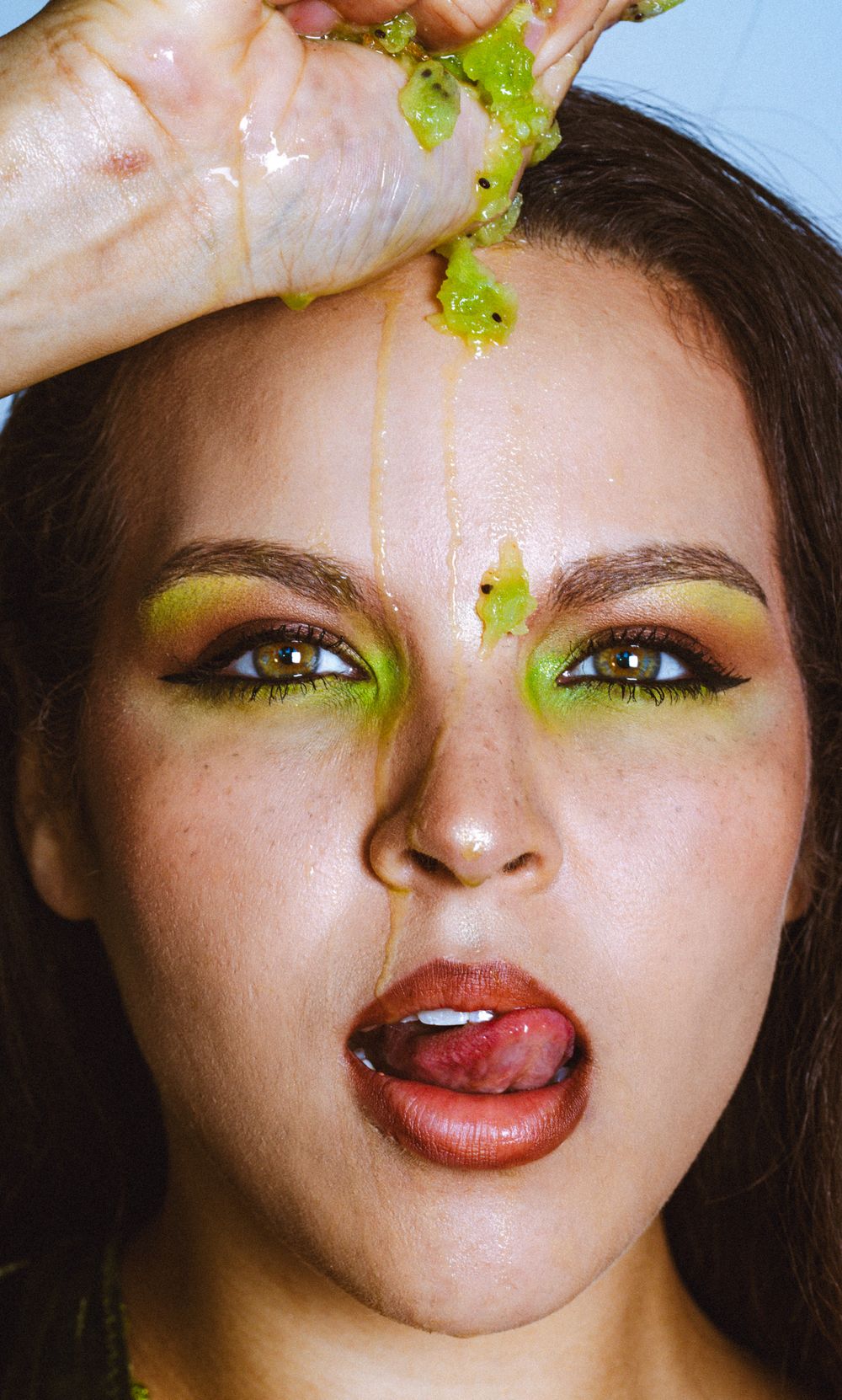 Woman with green eyeshadow and juice dripping down her face while squeezing a kiwi fruit above her head.