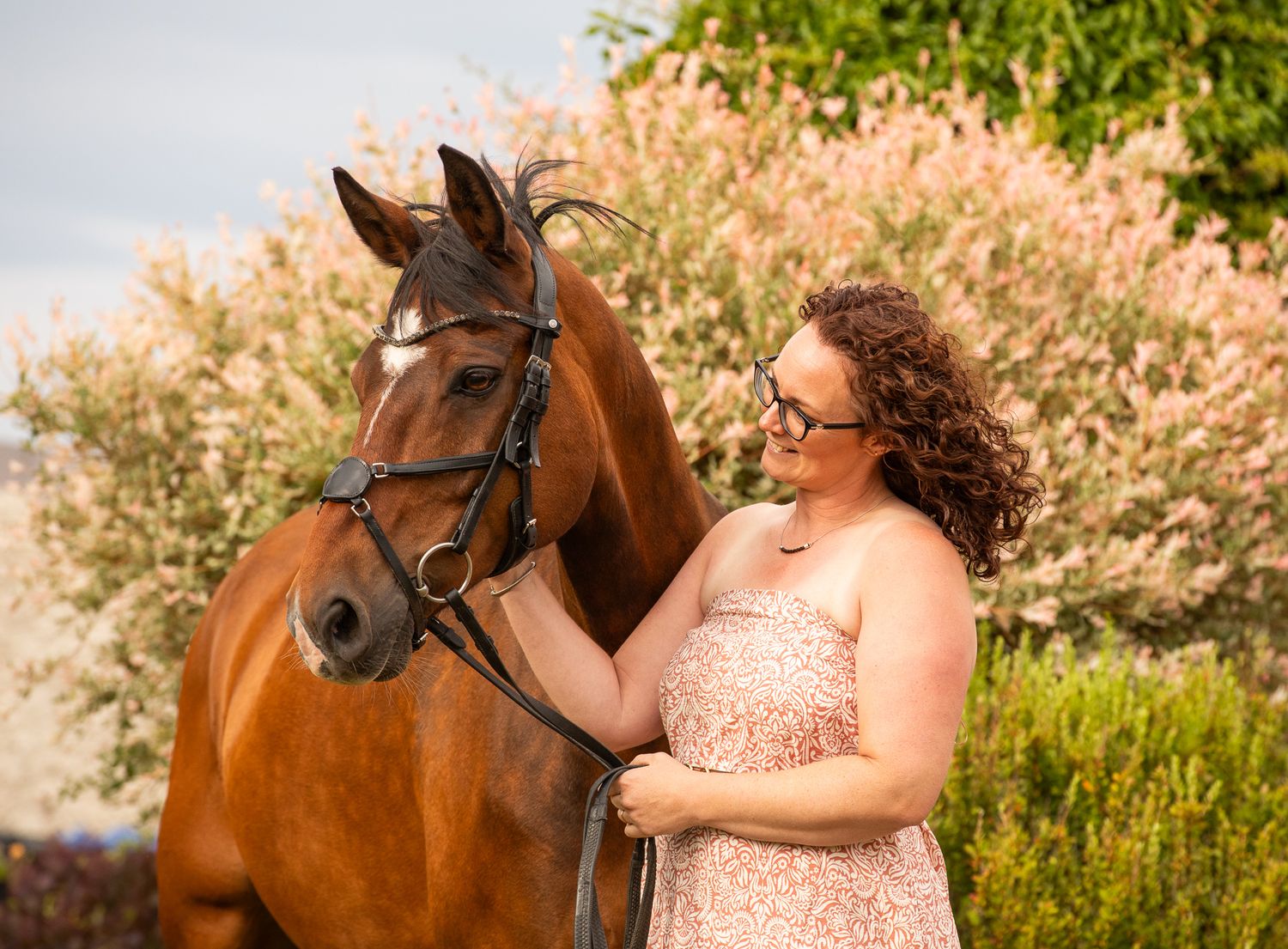 A person in a floral dress poses with a brown horse in a lush outdoor setting during golden hour.