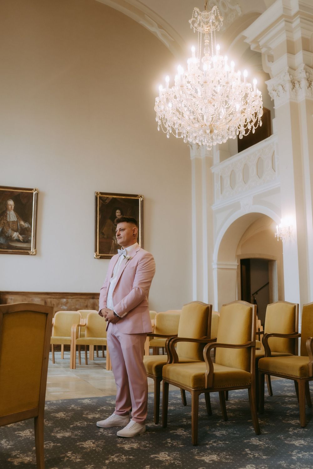 Grand reception room with crystal chandelier and person in pink suit standing near vintage chairs.