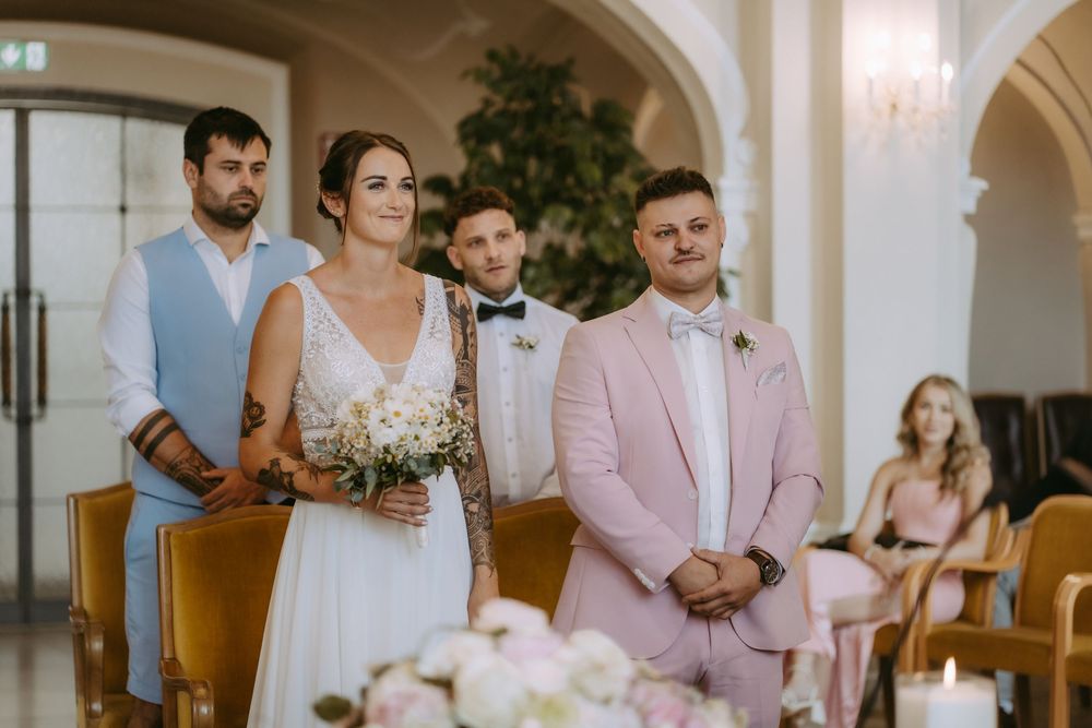 Wedding party gathered in elegant venue with arched windows and chandeliers.
