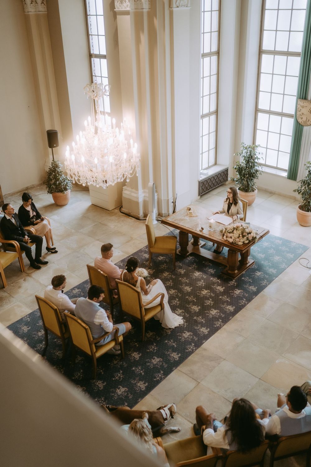 Elegant wedding ceremony in a bright white room with tall windows and crystal chandeliers.